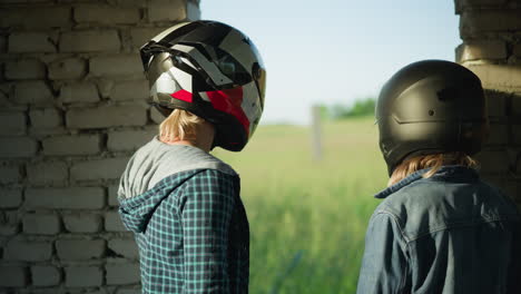 two women in helmets stand near the opening of an abandoned building, gazing toward a grassy field