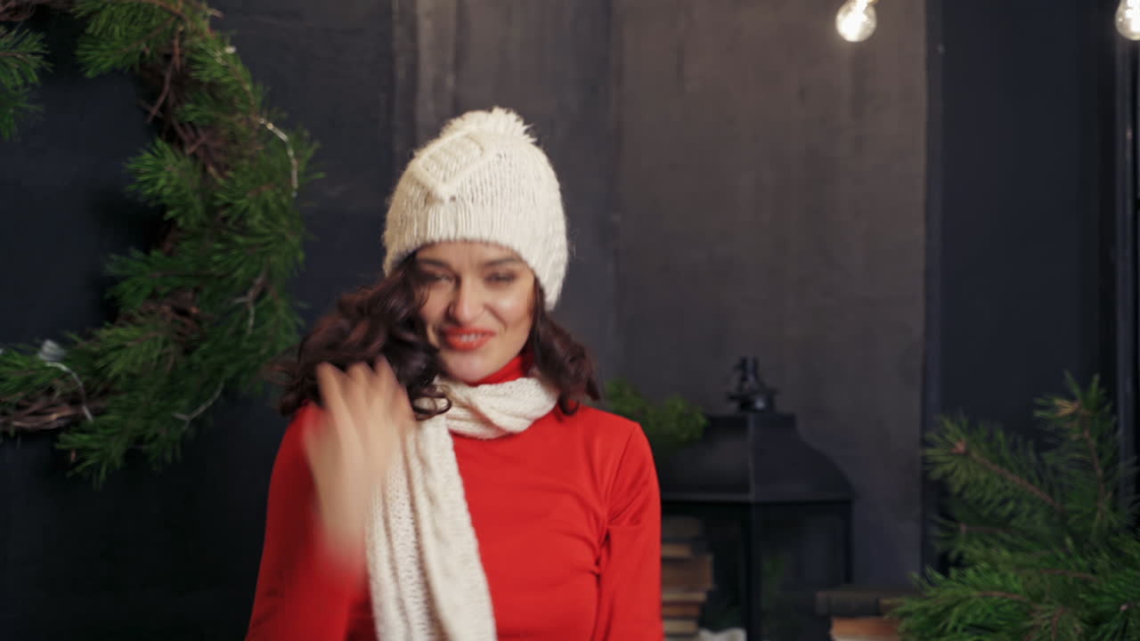 Lady putting on white knitted hat. Beautiful woman poses to the camera in room decorated before winter holidays. Fest spirit.