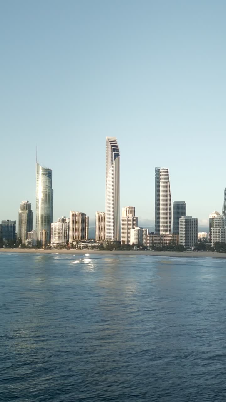 Vertical drone flying around revealing buildings with surfers in ocean, Gold Coast skyline, Queensland Australia. Dynamic aerial showcasing surf culture and luxury beachfront living at paradise