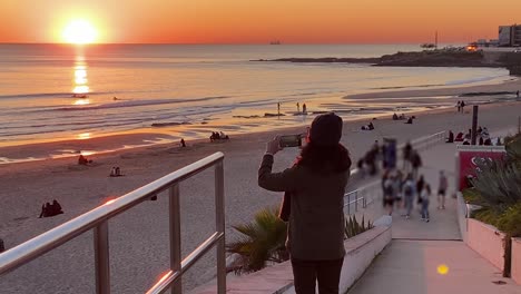 view of woman recording huge sunset with unrecognised people in background at carcavelos beach, small waves form and break, beautiful atlantic ocean, deep yellow sunset on water reflections, portugal