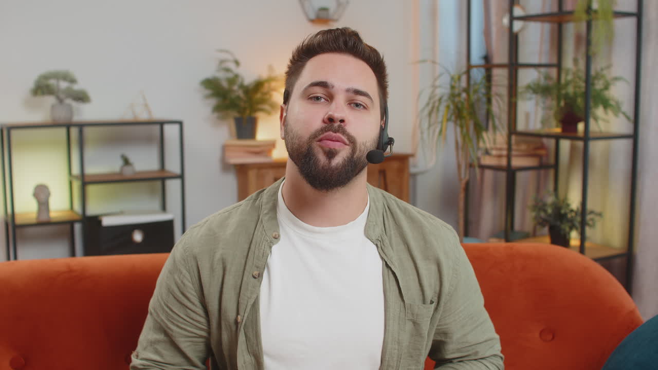 Confident young caucasian man guy sitting on sofa at home wearing headset talking with client