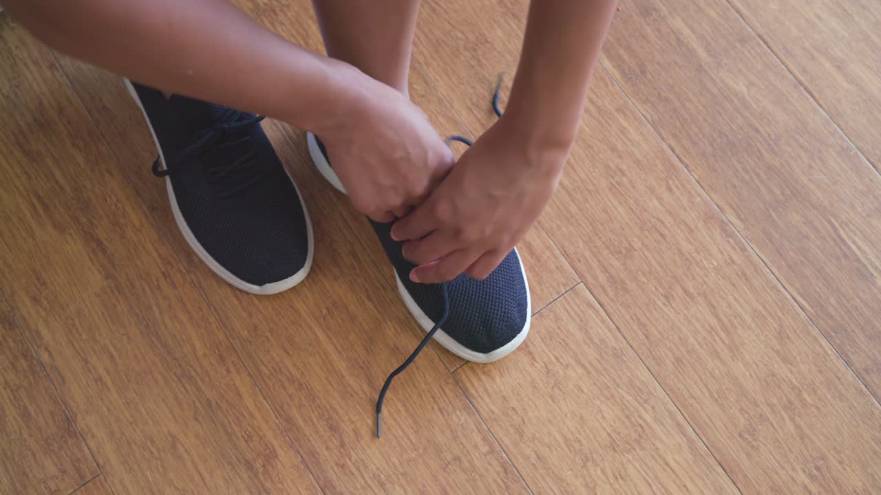 Una donna afroamericana in pantaloncini, seduta a casa, si allunga per allacciare i lacci delle scarpe da ginnastica, preparandosi ad alzarsi.