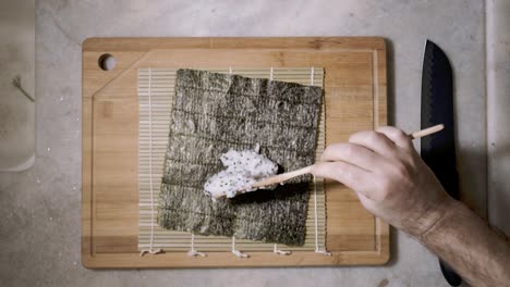 lapso de tiempo de arriba hacia abajo de dos manos con cuchara de madera y tableta de corte en el mostrador de la cocina esparciendo arroz con semillas de chai en una estera y hoja de nori de algas para hacer sushi