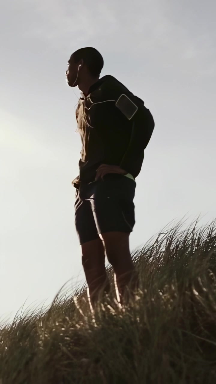 Video verticale: Uomo che raggiunge la cima, si ferma e osserva il panorama delle dune a metà corsa, con felpa, fascia da braccio e auricolari.