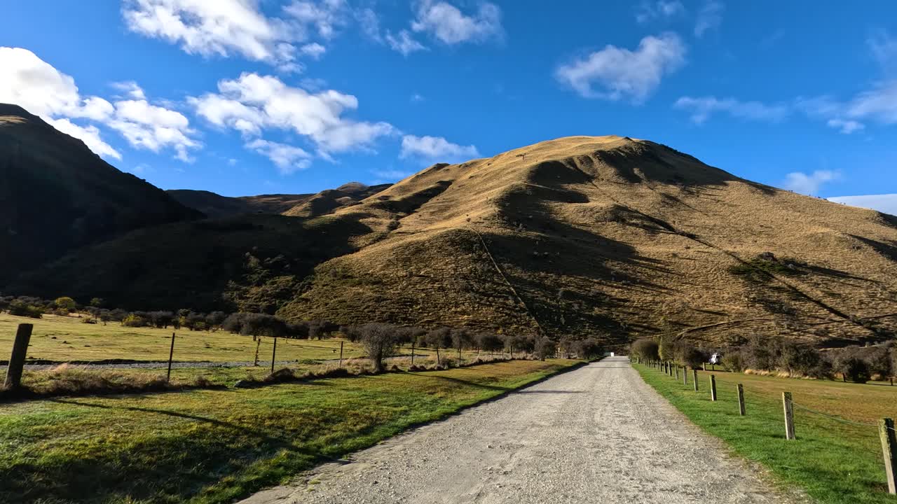Vehicle moves along rural gravel road toward sunlit mountain under blue sky, steady forward motion