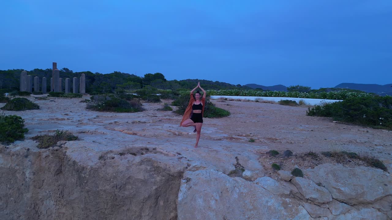 Woman performing a yoga tree pose on a rocky cliff during a serene sunset in Cala Llentia Ibiza. Smooth aerial view flight static tripod hovering drone