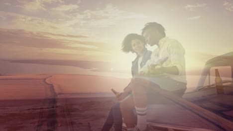 animation of african american couple sitting on car drinking beers at beach over sunset landscape