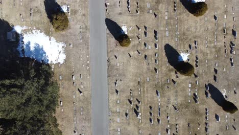 drone flying diagonally looking straight down at a cemetery with gravestones, dead grass, snow patches and trees that cast shadows on a sunny winter day