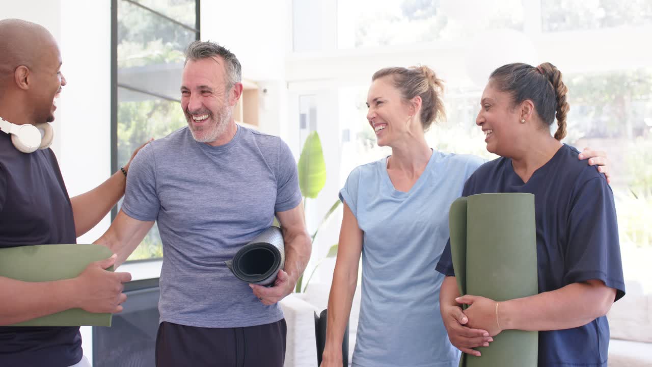Diverse Freunde mit Matten begrüßen sich vor dem Unterricht in einem sonnendurchfluteten Fitnessstudio, wobei der linke Mann eine Hand auf die Schulter legt.