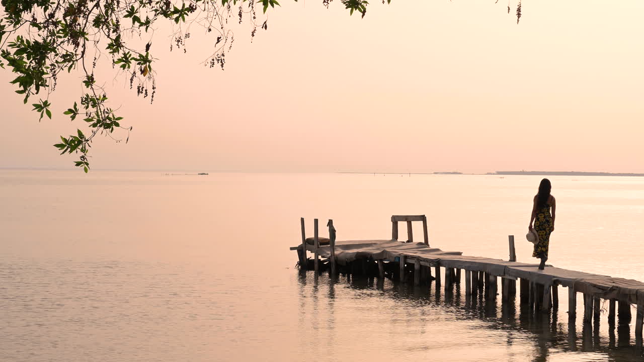 Woman walking to wooden pier relaxing on sea view water, with wind blowing on tree leaves at sunset over sky background