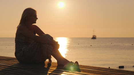 An-Adult-Woman-Sits-In-A-Summer-Sarafan-On-The-Beach-Sits-On-A-Bench-And-Holds-Onto-A-Suitcase-On-Wh