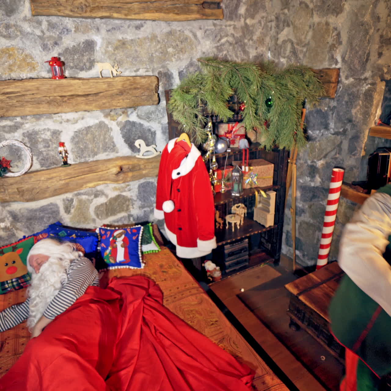 Santa Claus is sleeping in specially decorated room in his bed. Hand under cheek. Elves are ringing bell and touching him. Christmas spirit concept close up video. Video from above.