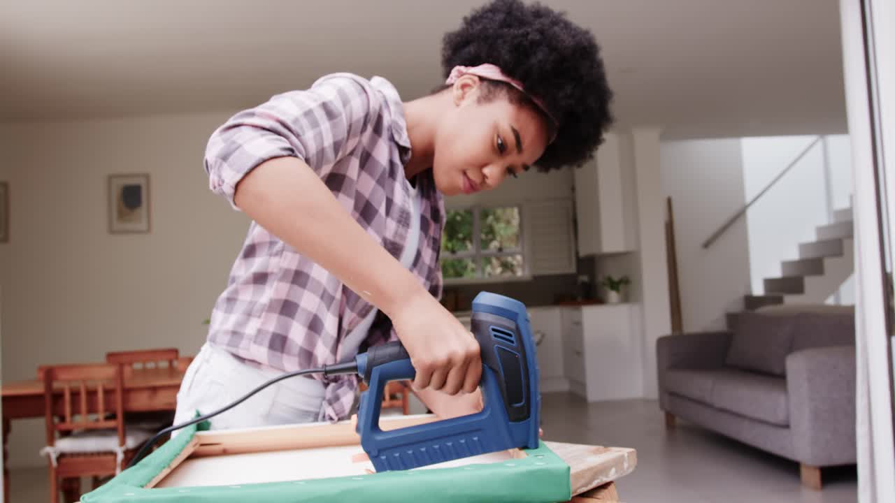 Donna afroamericana che afferra una cucitrice blu, chinata su una cornice e fissa un tessuto verde in casa.