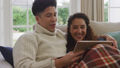 video of happy biracial couple siting on sofa under blanket and using tablet