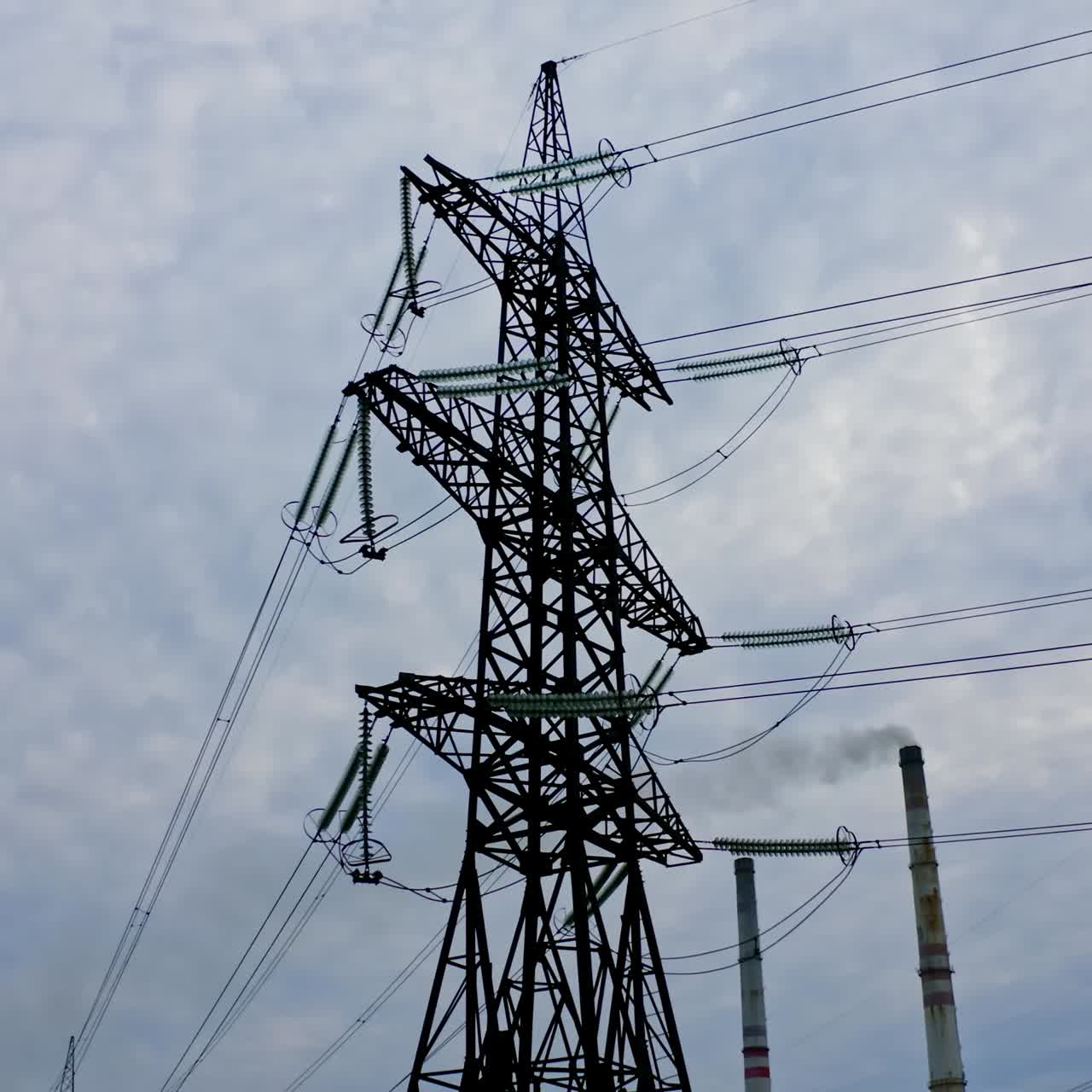 Electrical pylons near the industrial pipes. High voltage electrical lines on the evening sky background. Smoke from manufacture pollute the air.