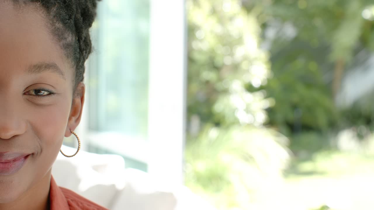 Adult woman of African descent smiling on sofa beside glass door, reacting to sunlight, copy space