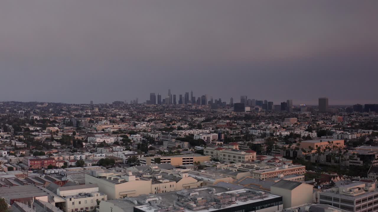 Low and wide aerial shot of downtown Los Angeles blanketed by smoke from the Eaton Fire during sunset in Southern California. 4K