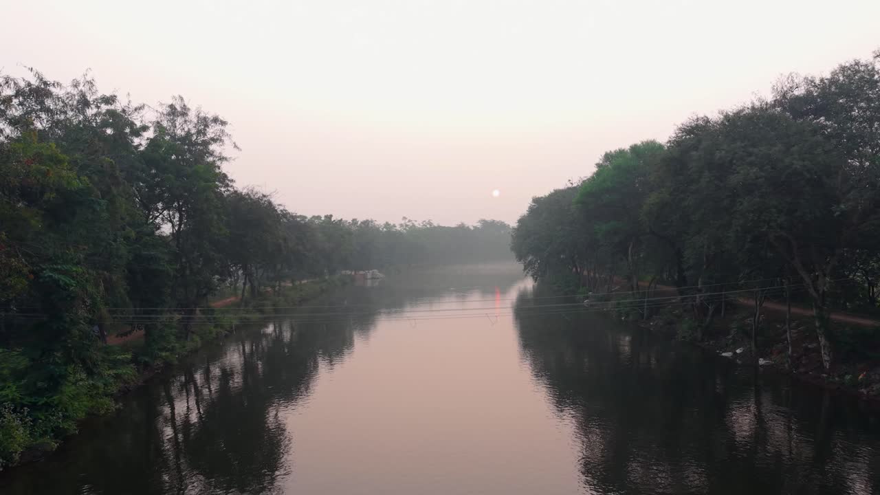 Fredelig kanal som flyter gjennom frodige, grønne trær ved soloppgang, og som reflekterer et mykt morgenlys på stille vann. Rolig landlig landskap med naturlig symmetri, mystisk atmosfære og et miljø preget av landsbygda