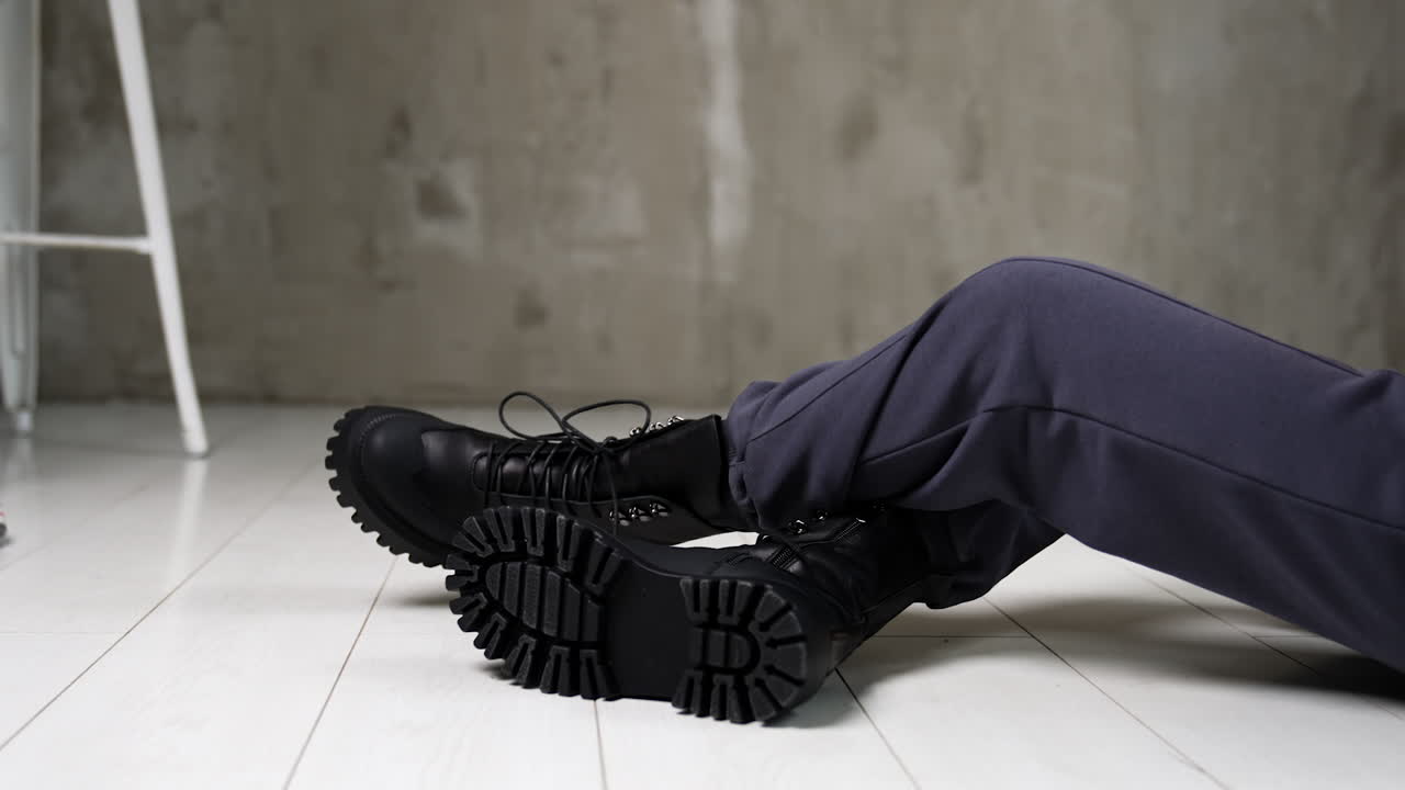 Model in grey sportive pants sitting on the white floor in studio. Female touching the tongues of black stylish army boots on her feet.