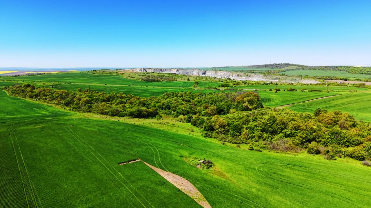 Clear day over green farmland near a quarry. Aerial view shows crop fields, forest and an exposed stone quarry on the horizon.