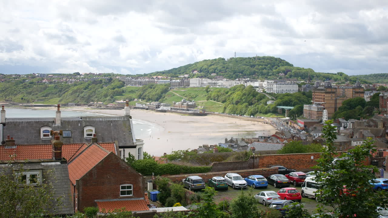 A coastal townscape in Scarborough, North Yorkshire, England shows rooftops, parked cars, and a sandy beach with hills and buildings in the background