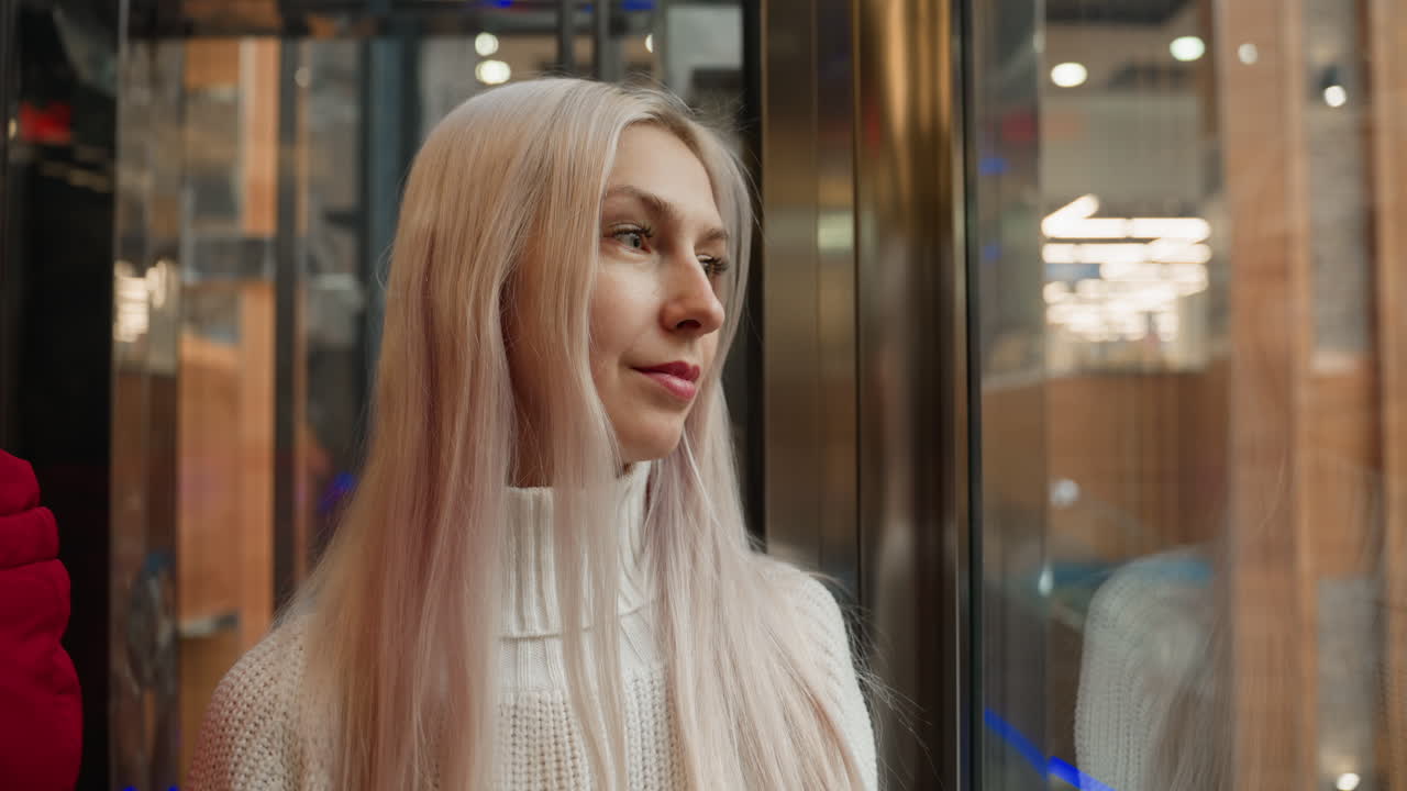 Shoulder view of elegant shopper standing inside elevator cabin, gazing thoughtfully through reflective glass, expression revealing introspection and calm anticipation amid upscale retail environment