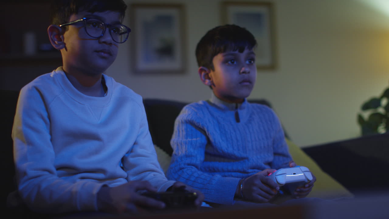 Two Young Boys Sitting On Sofa At Home Playing With Computer Games Console On TV Holding Controllers Late At Night 5