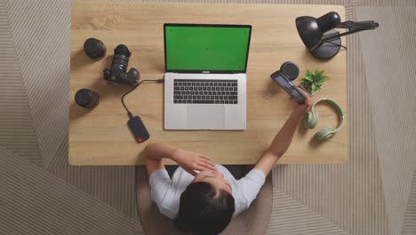 top view of a woman video editor waving hand and having a video call on smartphone while using a green screen laptop next to the camera in the workspace at home