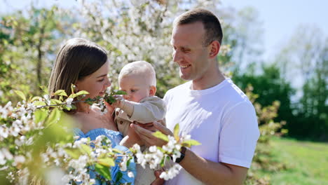 happy young parents hold their cute blond baby son standing in the spring garden. serious little kid touches the tree branch.