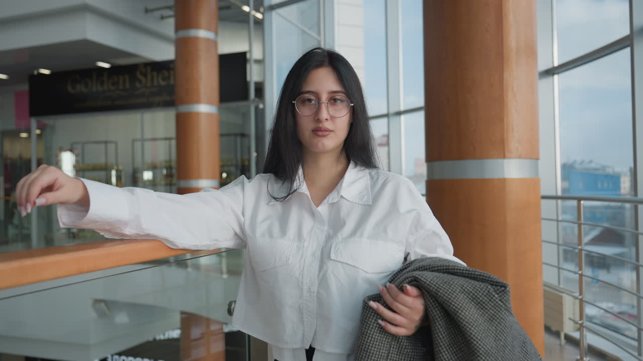 Elegant woman in white shirt confidently adjusting glasses while standing beside indoor pillar in modern commercial space with glass walls and daylight filtering through large windows