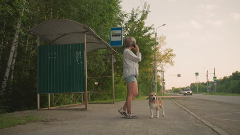 woman standing at bus stop in rural area holding dog on leash while talking on phone, surroundings include green trees, bus stop shelter, and road with cars passing by