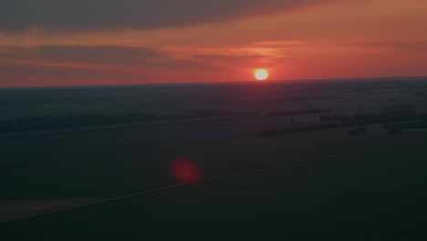 blazing red sun setting over distant farmland horizon casts warm orange glow across fields and sky while subtle lens flare accentuates tranquil dusk atmosphere in sweeping aerial sunset panorama