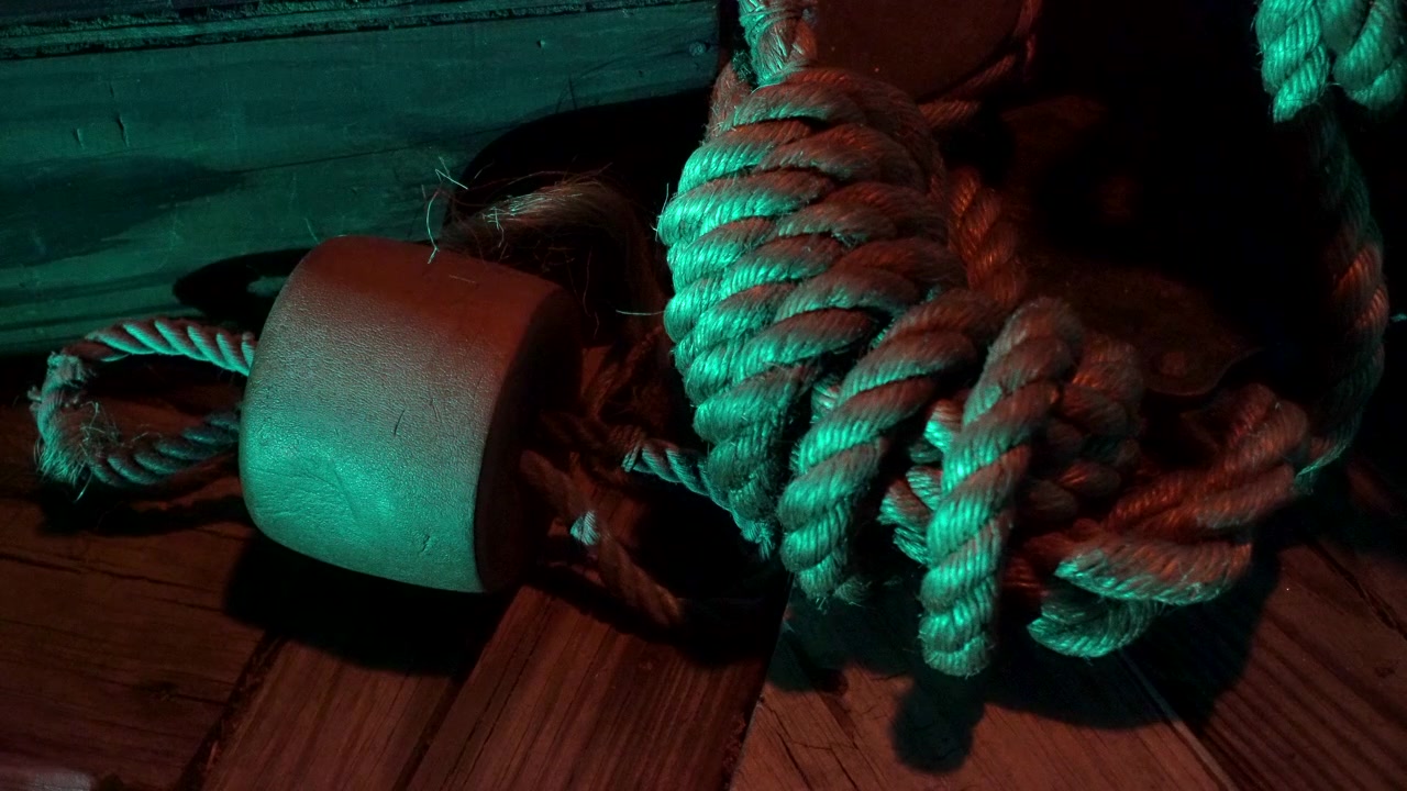 Corda enrolada e uma boia de cortiça sobre um deque de madeira iluminado por tochas.