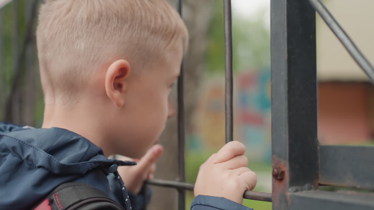 Young boy at school gate ready with backpack, navy jacket and steady grip on metal bars, hopeful gaze into campus, preparing to go back after break, clear daylight, closeup hands and profile, composed