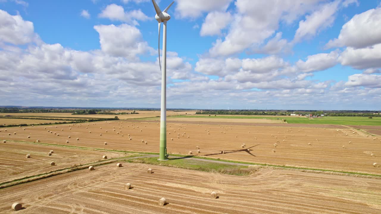 The aerial video captures the tranquil scene of wind turbines spinning in a Lincolnshire farmer's freshly harvested field, where the golden tones of hay bales add charm