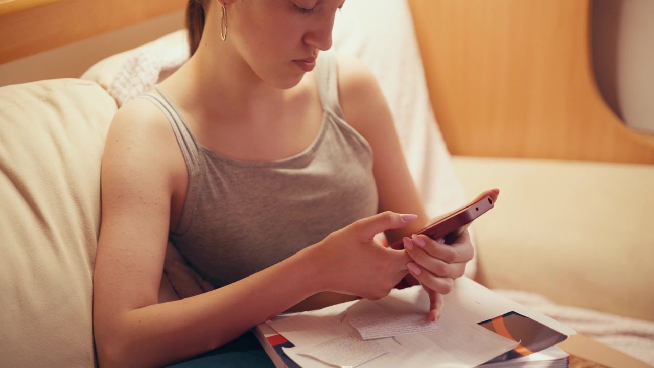 Focused high school girl sitting on a couch and typing on her mobile phone, using a smartphone to copy notes, browse exam answers, or cheat on a future test while studying at home.