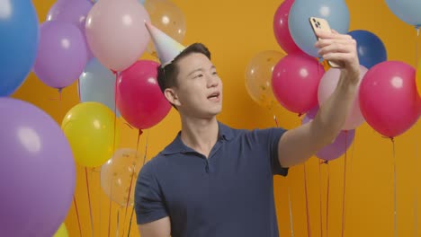 studio portrait of man taking selfie wearing party hat celebrating birthday surrounded by balloons 1