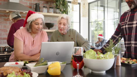 happy diverse senior female friends preparing christmas meal using laptop in kitchen, slow motion