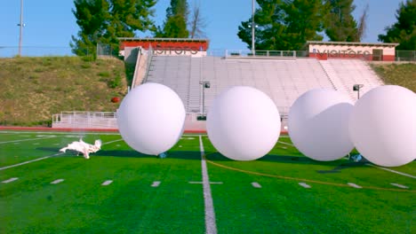 big white balloons filled with air being crushed by an american football on an american football field in slow motion during summer time while sun shines