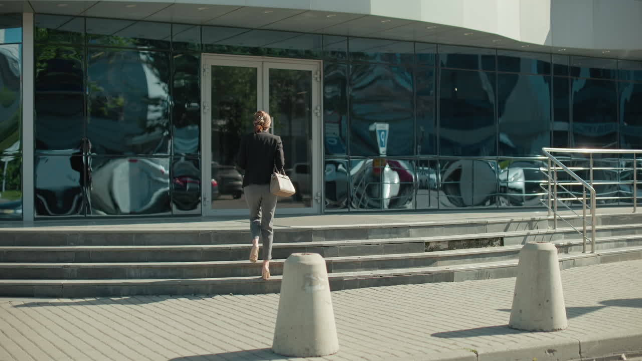 Businesswoman walking towards modern glass office building holding handbag, with sleek reflections of parked cars on facade, showcasing professional lifestyle in urban setting