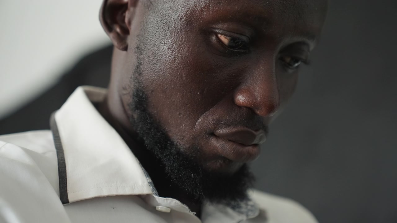 Primo piano di un uomo stanco che guarda in basso, fatica post-turno in un interno fioco, barba sottile e colletto, sudore sulla fronte, grana cinematografica, riflessione silenziosa, messa a fuoco superficiale, ombre cupe, stanco.