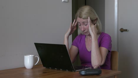 mujer con anteojos con dolor de cabeza por migraña trabajando en una laptop, con exceso de trabajo en casa