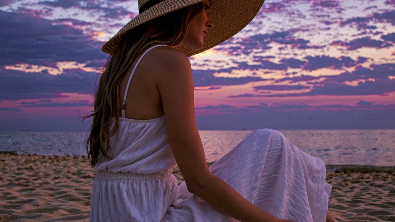 A woman sits peacefully on the warm sand of a beach as the sun sets, casting a beautiful glow across the horizon. She wears a wide-brimmed sun hat and a flowing white dress, perfectly complementing the vibrant colors of the sky. The tranquil atmosphere is enhanced by the gentle waves lapping at the 