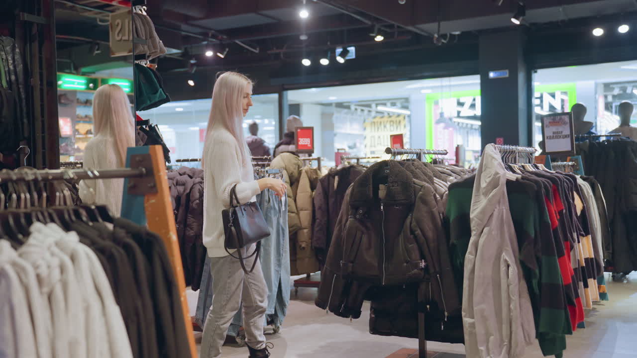 Fashionable shopper with long silky hair and black handbag walks through clothing boutique browsing coat rack filled with winter jackets and colorful sweaters under indoor lighting