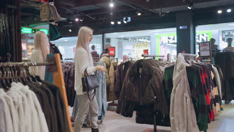 fashionable shopper with long silky hair and black handbag walks through clothing boutique browsing coat rack filled with winter jackets and colorful sweaters under indoor lighting