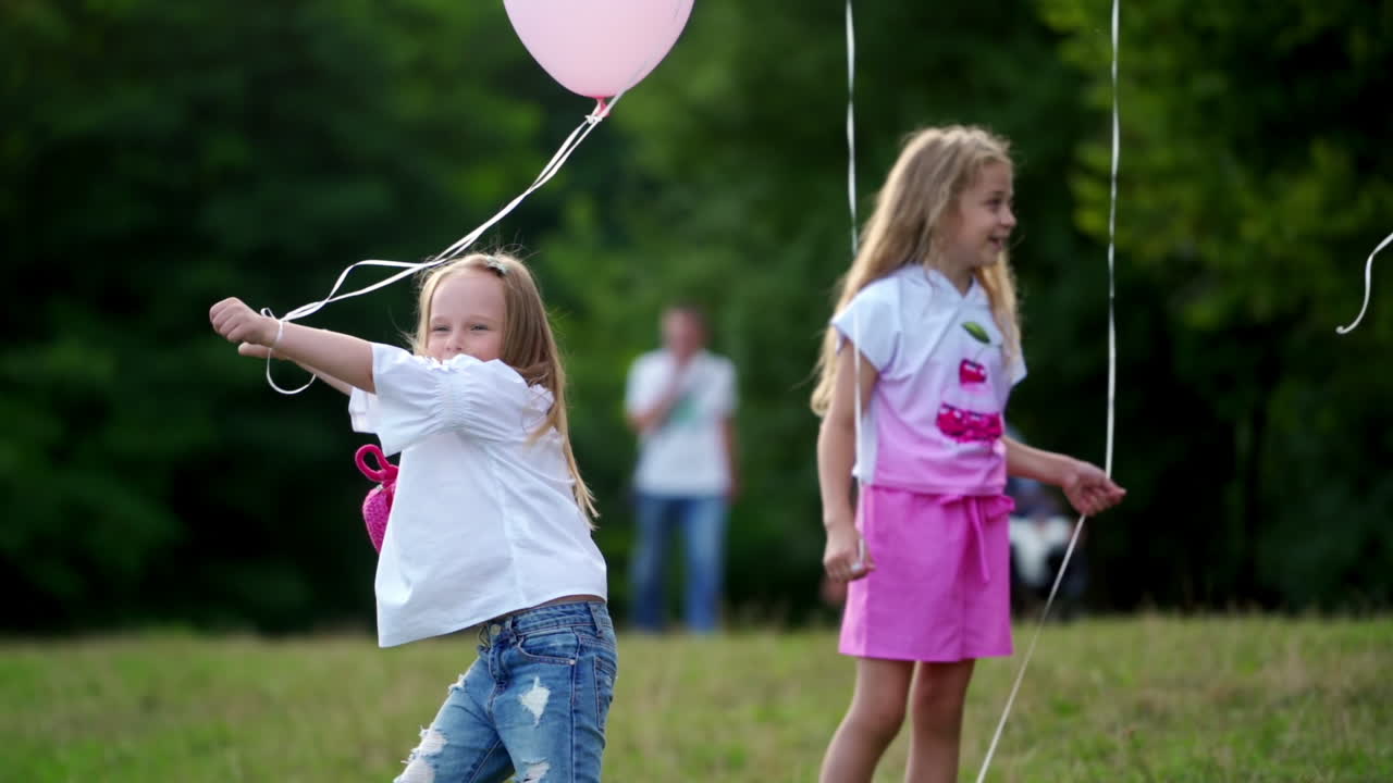 Little girls playing with baloons outside. Cute sunny kids dance with baloons on green grass. Happy holidays outside in summer day. One girl is standing nearby and smiling.