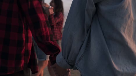 Close-up-footage-of-a-couple-holding-hands-together-while-walking-on-the-beach.-Another-couple-on-a-perspective.-Young-people-having-fun-together.-Dusk