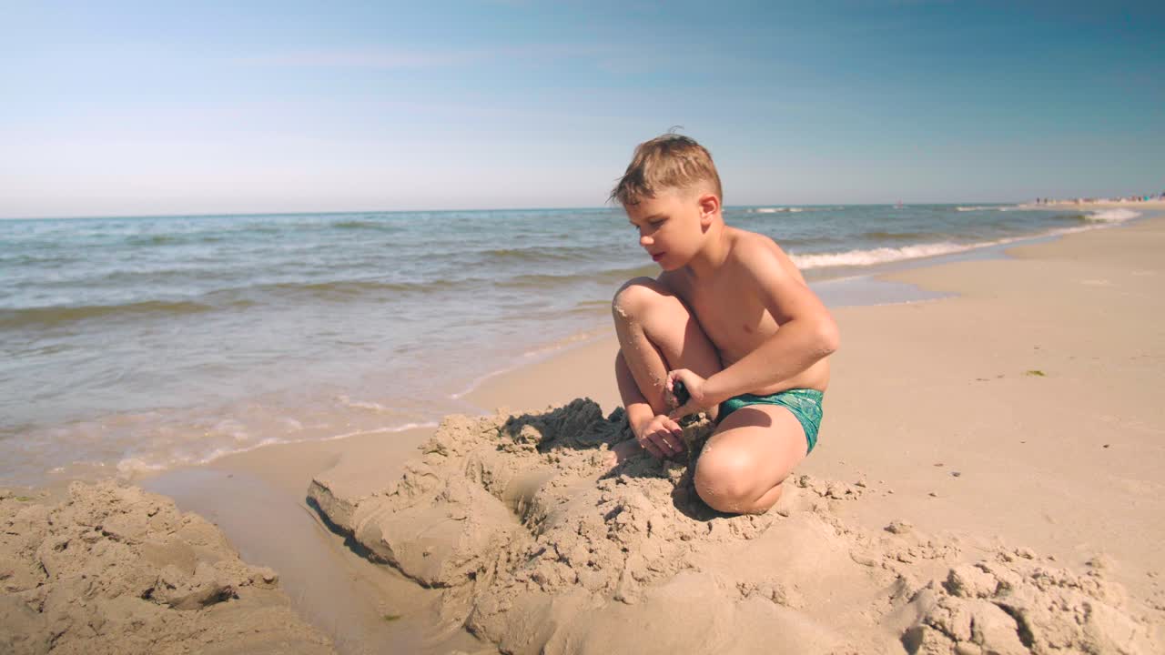 Young boy at the beach is busy creating a path into the sand next to shoreline