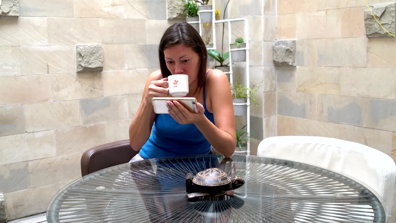 Woman drinking tea and watching funny videos on the phone while sitting at a table