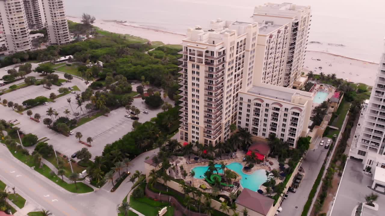 A drone semi-orbit shot circles a coastal city at sunrise, showcasing modern skyscrapers, ocean waves, and a golden sky from multiple angles.